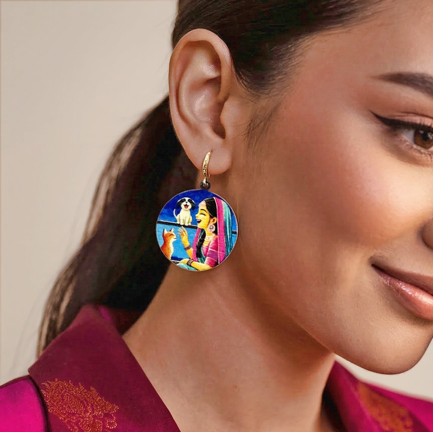 A close-up of a woman wearing a colorful round earring with a gold-tone hoop. The earring features a vibrant illustration of a woman and two dogs under a blue sky, styled with a magenta top against a soft beige background.